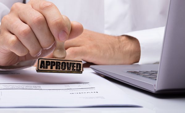 Close-up Of A Person's Hand Stamping With Approved Stamp On Document At Desk